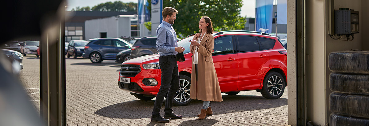 Zwei Personen sprechen am Autohaus, ein rotes Auto im Hintergrund, sonniger Tag.